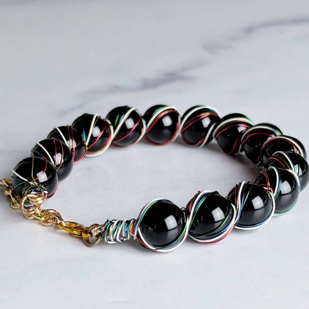 A beaded bracelet featuring glossy black round beads wrapped with thin silver, red, and green metallic wires, finished with a gold chain clasp, shown on a light gray marble surface.