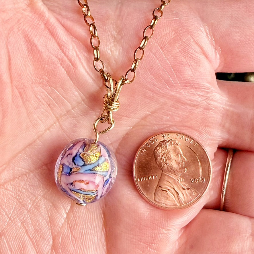 A close-up of a handmade glass bead pendant necklace with a gold chain, held in a person's hand next to a US penny for scale. The pendant is a round glass bead featuring a marbled swirl pattern of purple, pink, blue, and gold foil.