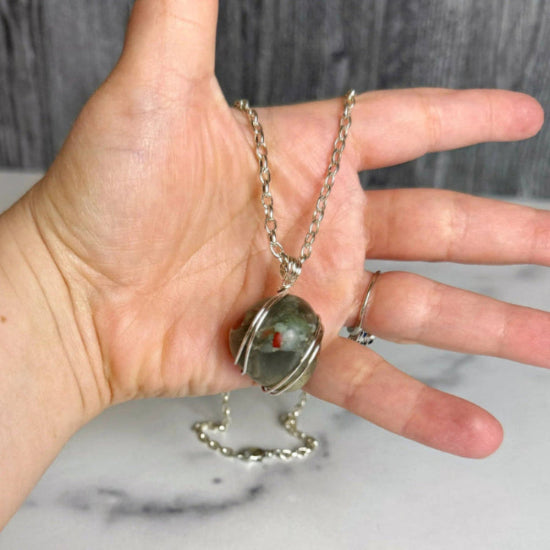 Hand holding a necklace with a bloodstone pendant against a neutral background