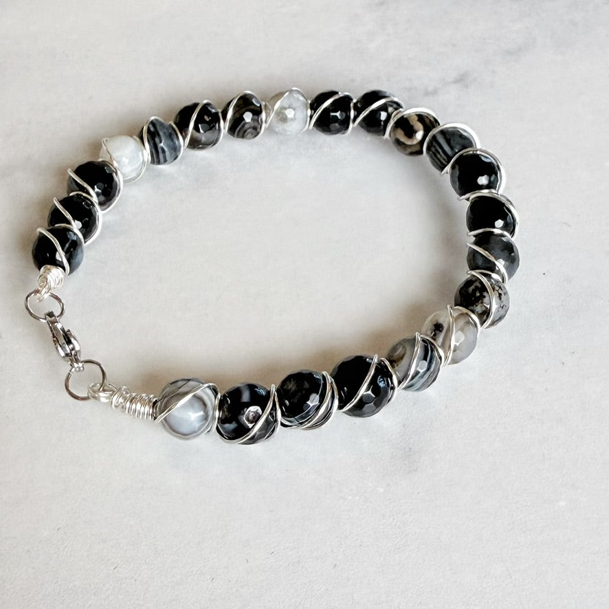 A beaded stretch bracelet featuring black and gray agate beads, a white pearl, and silver wire-wrapped accents, displayed on a light gray marble surface.