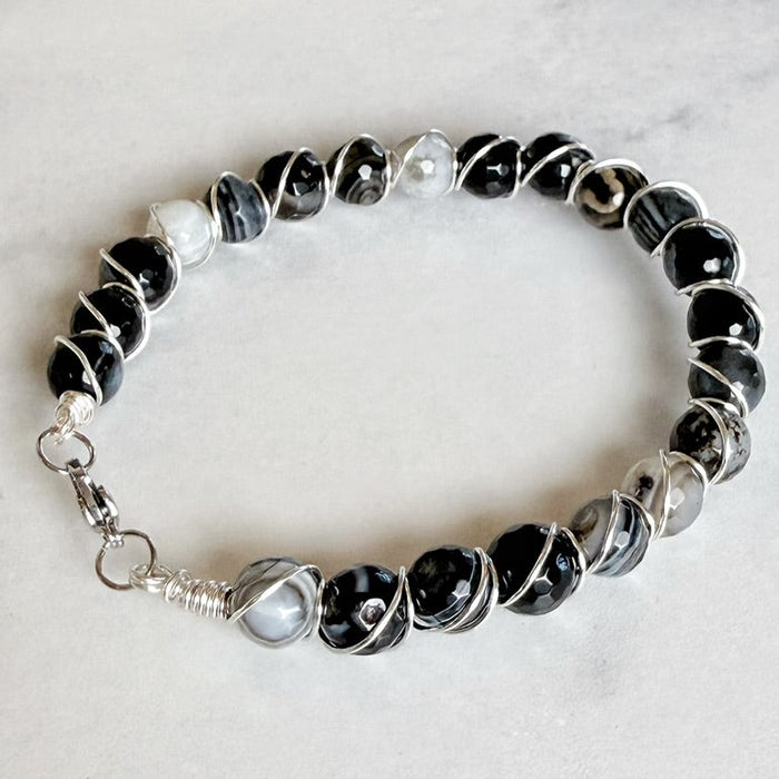 A beaded stretch bracelet featuring black and gray agate beads, a white pearl, and silver wire-wrapped accents, displayed on a light gray marble surface.