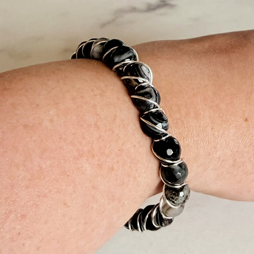 A close-up of a beaded stretch bracelet worn on a person's wrist. The bracelet features glossy black and gray stone beads separated by twisted silver metal bands.