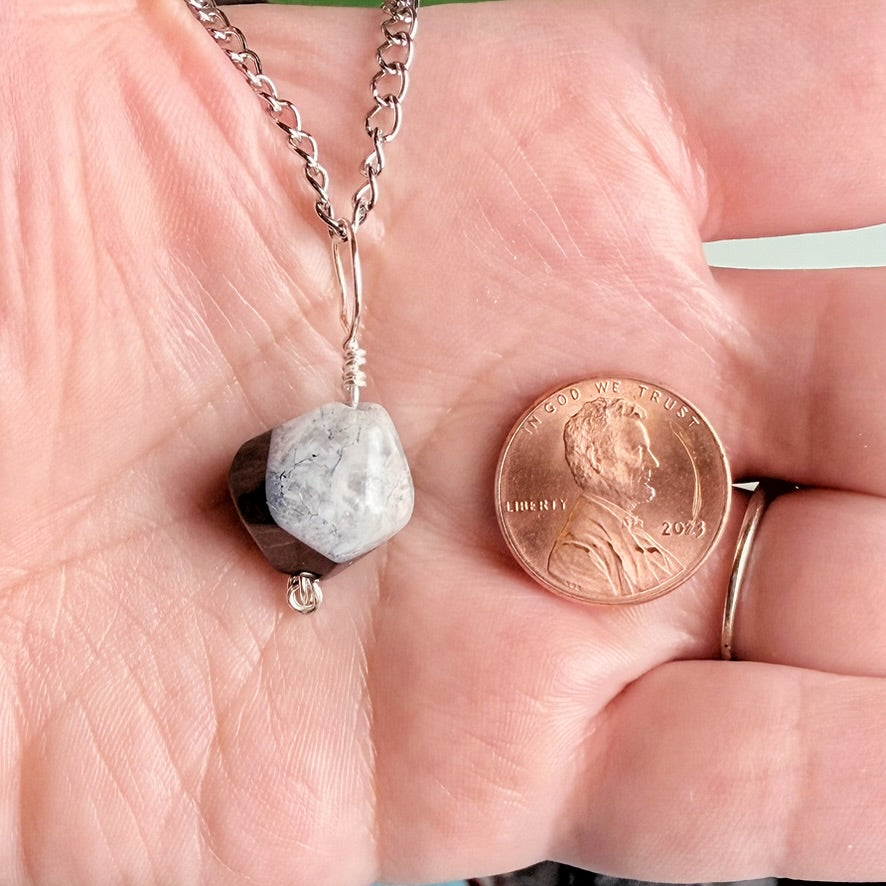 A silver chain necklace with a rough, dual-toned stone pendant (one half light gray, the other half dark brown/black) held in a person's palm next to a 2013 US penny for scale.