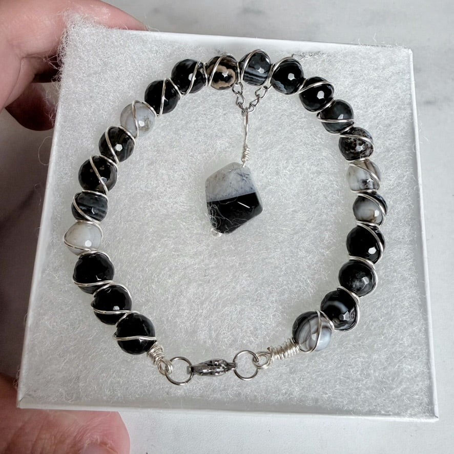 A beaded bracelet displayed on a white jewelry box, featuring black and white marbled beads, silver wire-wrapped accents, and a central irregular black-and-white stone charm. A hand is holding the box.