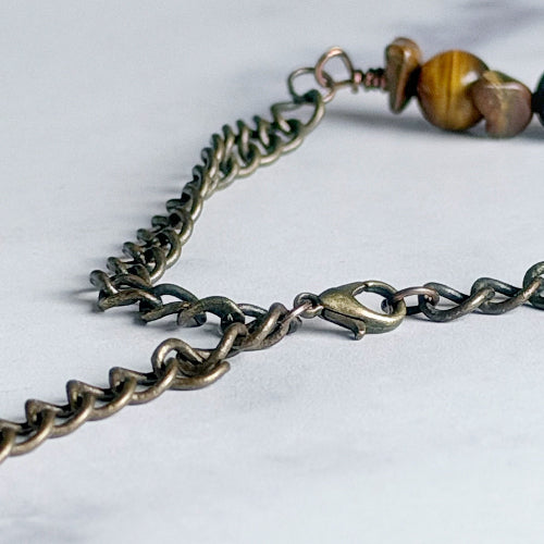 Close-up of a dark bronze metal chain necklace with a lobster clasp and a section of brown and gold tiger's eye beads on a light gray background.