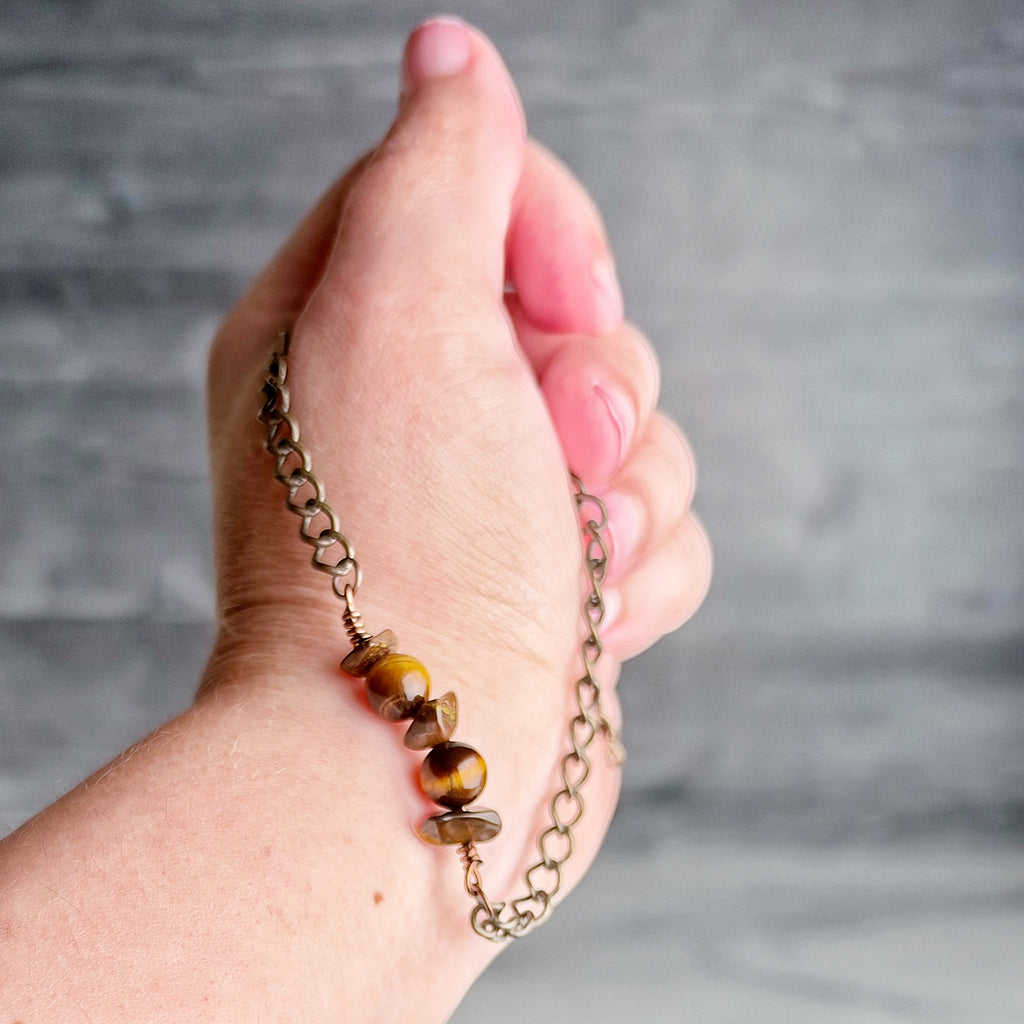A close-up of a hand holding a delicate chain bracelet with three round tiger's eye beads and small metallic spacer pieces, set against a gray textured background.