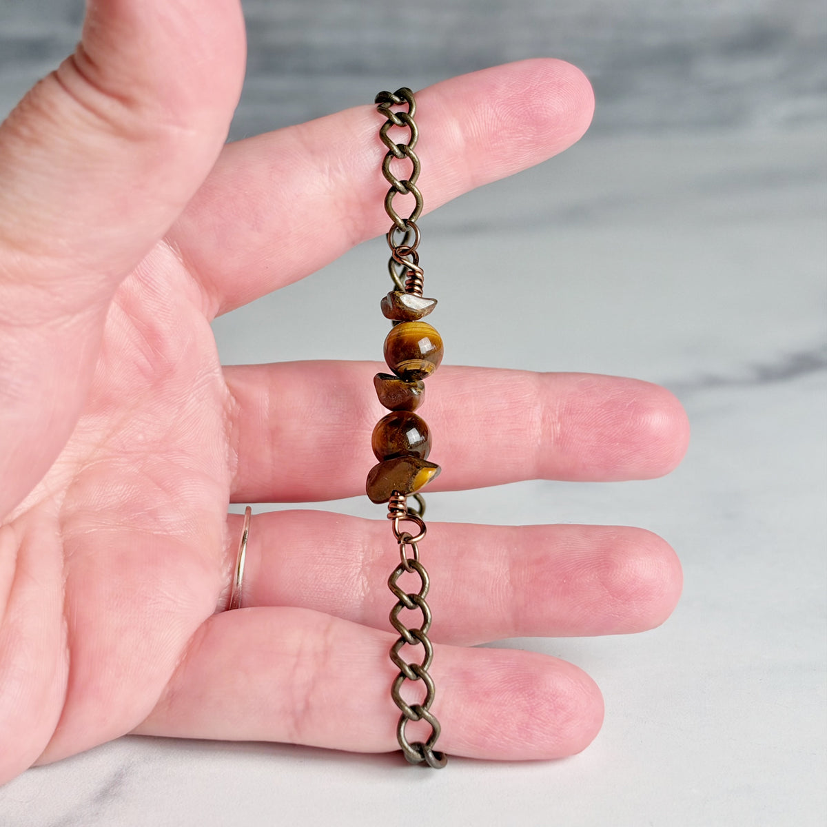 A close-up of a hand holding a short chain bracelet with polished tiger's eye beads and an antique bronze link chain against a light marble background.