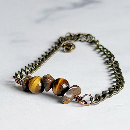 A handmade chain bracelet featuring a bronze-toned metal link chain, a twisted wire accent, and four polished tiger's eye beads in brown and gold tones, displayed on a light marble surface.