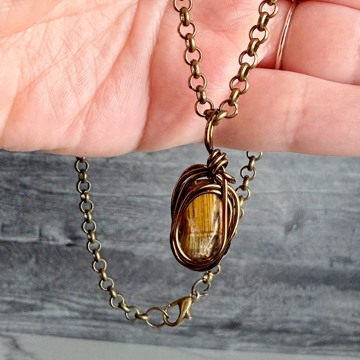 A close-up of a hand holding a bronze chain necklace with a tiger's eye pendant. The pendant features a polished golden-brown stone wrapped in textured bronze wire, set against a gray wood-grain background.