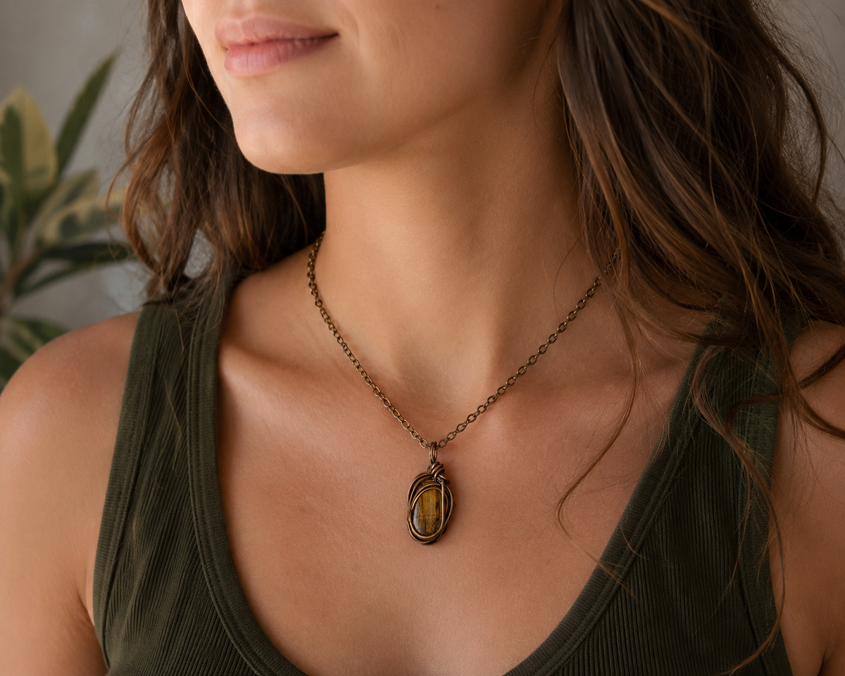 A close-up of a woman wearing a delicate bronze chain necklace with an oval pendant featuring a yellow and brown textured stone, paired with an olive green ribbed tank top.