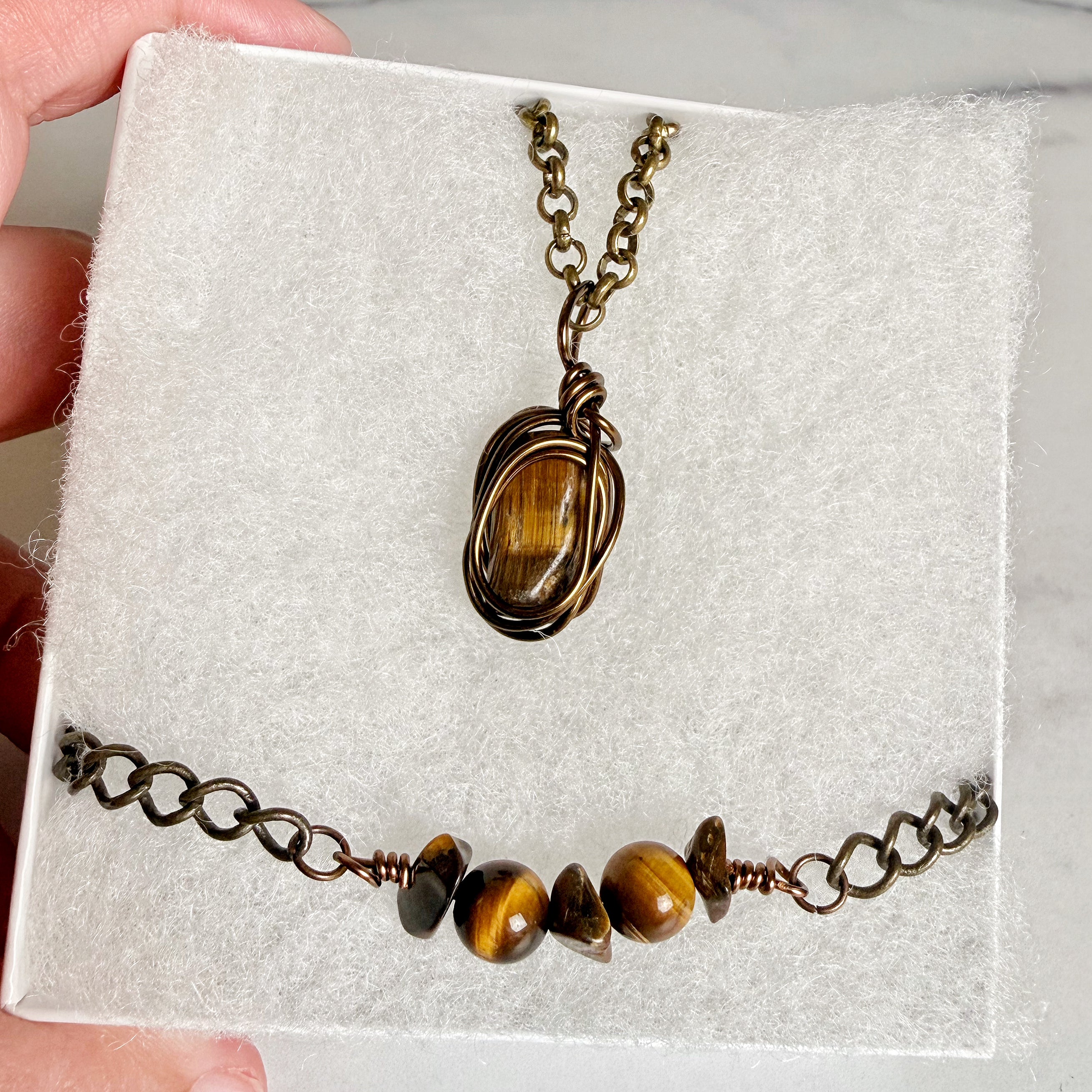 A jewelry set featuring a wire-wrapped pendant necklace and a beaded chain bracelet, both with tiger's eye stones, displayed on a white textured insert held by a hand.