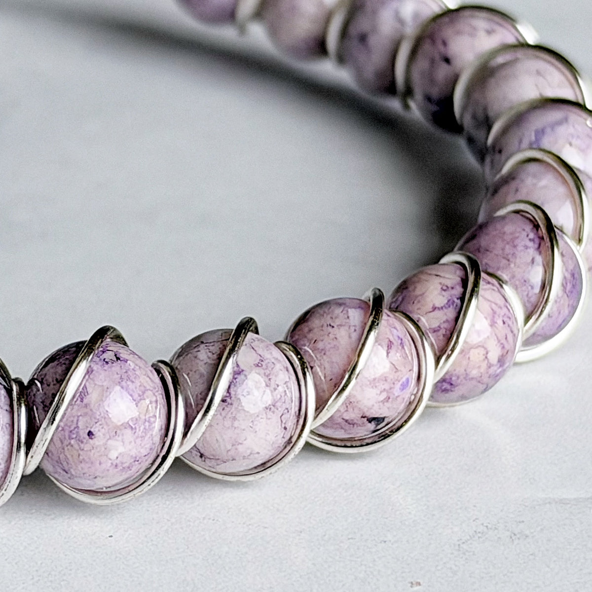 A close-up of a beaded bracelet featuring round lavender and purple agate beads wrapped in silver wire, set against a neutral light gray background.