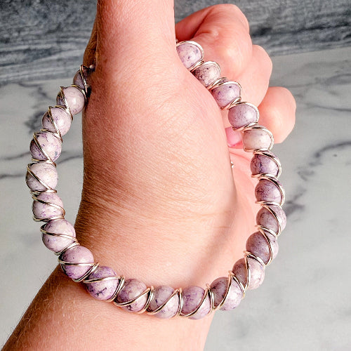 A close-up of a hand wearing a beaded bracelet made of round lavender-purple stones wrapped in silver-toned metal, set against a light marble background.