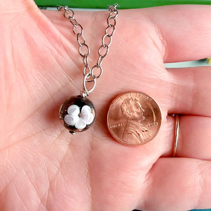 A silver chain necklace with a small round pendant containing white flower-like beads, shown resting on a person's palm next to a penny for scale.