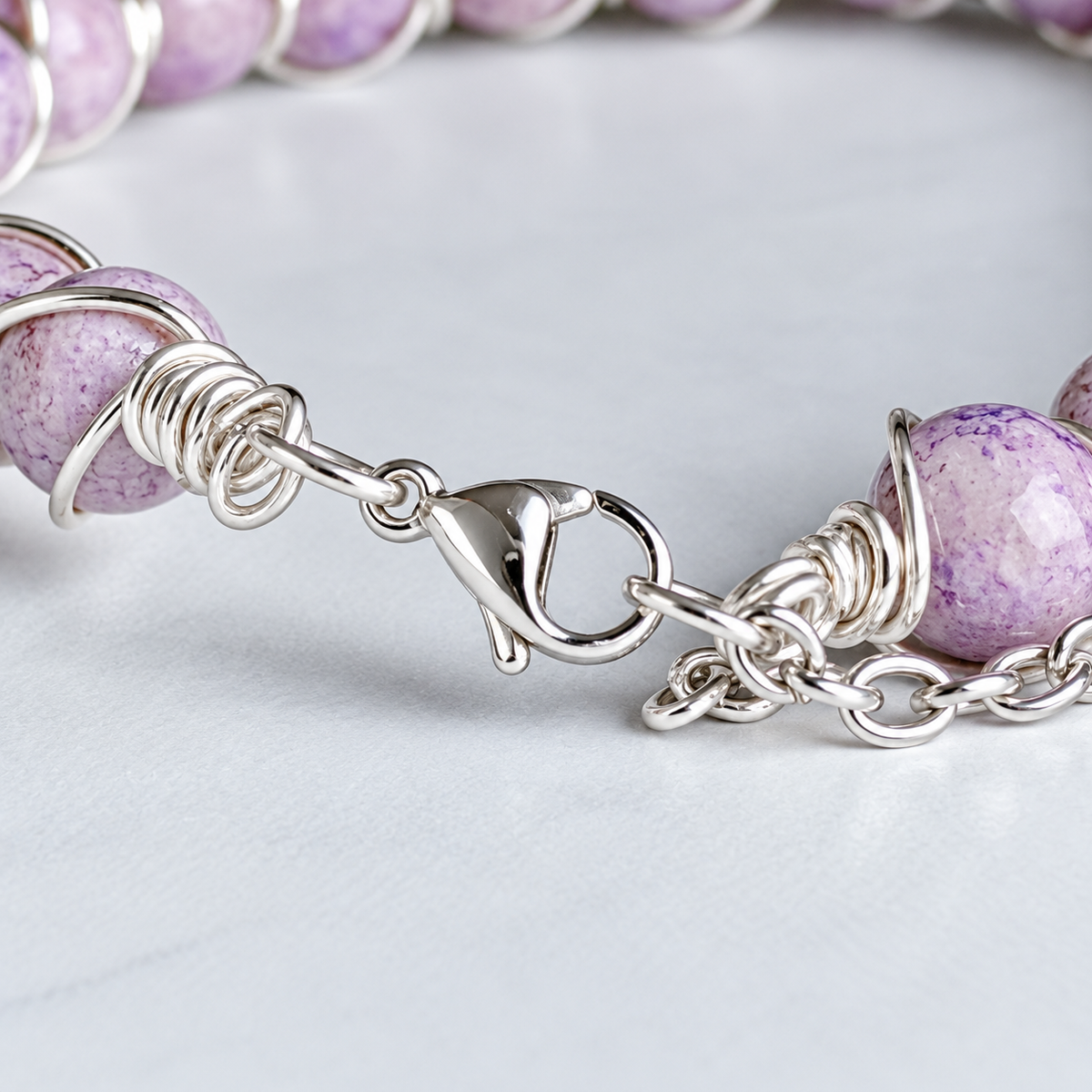 Close-up of a beaded bracelet with round lavender-purple marbled beads wrapped in silver wire and connected by a silver lobster clasp and chain, on a light gray surface.