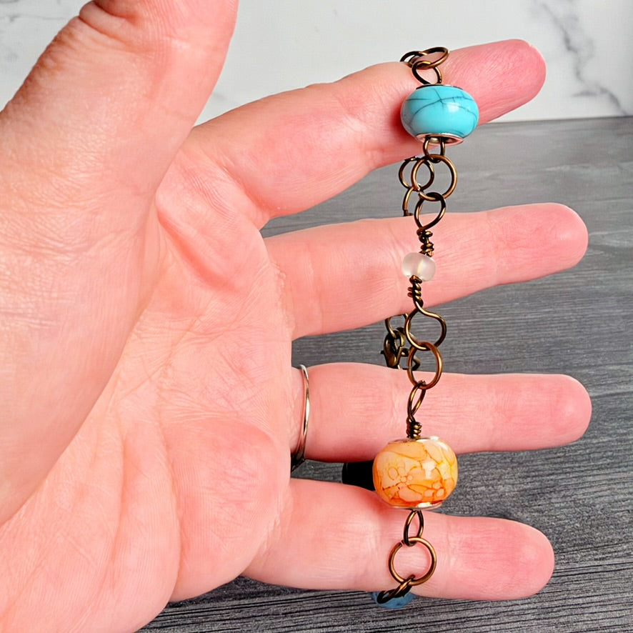A handmade charm bracelet with a turquoise bead, a marbled orange bead, and antique bronze metal rings, held in a person's hand against a gray wood background.