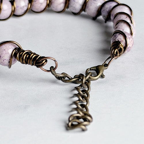A close-up of a beaded bracelet with pale lavender and white marbled beads wrapped in bronze-toned metal, featuring a chain clasp with a lobster clasp on a light gray background.