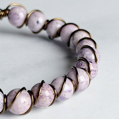 A close-up of a beaded bracelet featuring round, polished lavender and white stone beads wrapped in textured bronze wire, set against a light gray background.